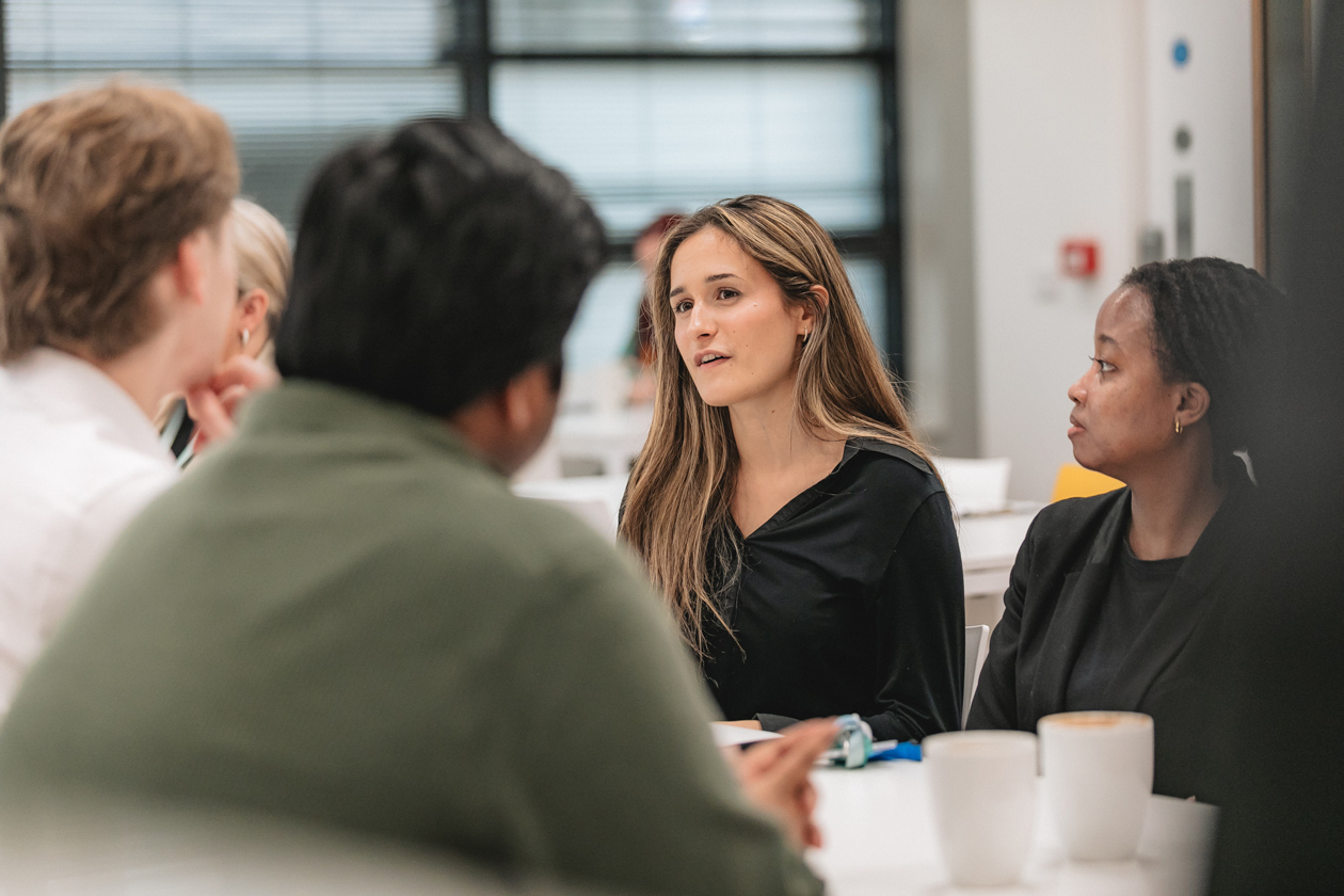 A lady speaking in front of two men on a table and another lady looking at her