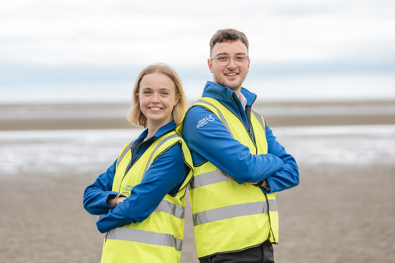 Two graduates folding their arms wearing a Uisce Éireann jacket with a high visible jacket on a beach 