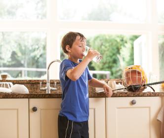 A boy drinking a glass of water in the kitchen with a hurl and helmet on the counter