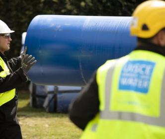 two uisce eireann workers caring a blue pipe over a glass plain with trees in the background 