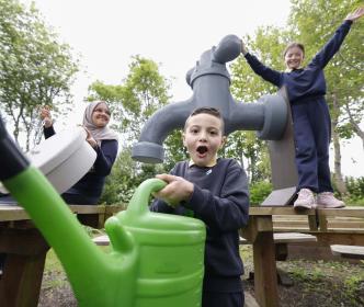 Three kids next to an oversized tap with one of them holding a watering can