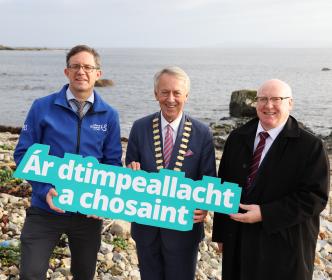 Three men holding a sign reading "Ár dtimpeallacht a chosaint" on a beach