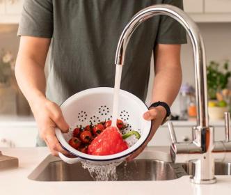 A man washing tomatoes and a red pepper in a colander with water from a tap